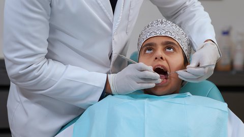 تصوير مقرب لولد عربي خليجي عماني يرتدي الزي التقليدي يجلس في العيادة السنية ويفتح فمه لتشخيص المرض ، معالجة النخر والتسوس,الحصول على ابتسامة مثالية، ايدي طبيب عربي خليجي عماني يرتدي القفازات الطبية يقوم بفحص أسنان المريض، الادوات اللازمة لإجراء الفحوصات