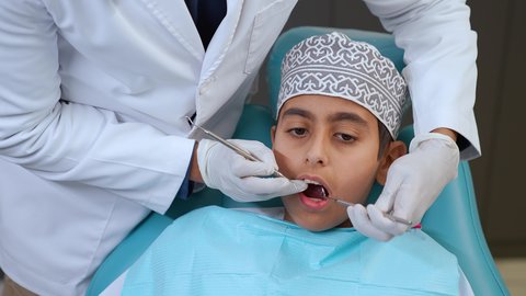 معالجة النخر والتسوس,الحصول على ابتسامة مثالية، ايدي طبيب عربي خليجي عماني يرتدي القفازات الطبية يقوم بفحص أسنان المريض، الادوات اللازمة لإجراء الفحوصات، تصوير مقرب لولد عربي خليجي عماني يرتدي الزي التقليدي يجلس في العيادة السنية ويفتح فمه لتشخيص المرض