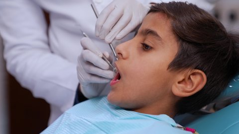 الادوات اللازمة لإجراء الفحوصات، معالجة النخر والتسوس,الحصول على ابتسامة مثالية، تصوير مقرب لولد عربي خليجي عماني يرتدي الزي التقليدي يجلس في العيادة السنية ويفتح فمه لتشخيص المرض, ايدي طبيب عربي خليجي عماني يرتدي القفازات الطبية يقوم بفحص أسنان المريض