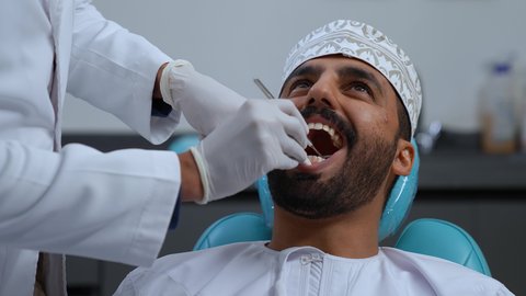 تصوير مقرب لطبيب يرتدي القفازات الطبية يقوم بفحص أسنان المريض وتشخيص المرض, العناية بالصحة الفموية، شاب عربي خليجي عماني يرتدي الزي التقليدي يجلس على كرسي العلاج، زيارة العيادة السنية لإجراء الفحوصات الدورية، تركيب الحشوات ومعالجة التسوس، المعدات اللازمة لإجراء الفحوصات الطبية