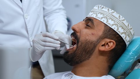 العناية بالصحة الفموية، شاب عربي خليجي عماني يرتدي الزي التقليدي يجلس على كرسي العلاج، زيارة العيادة السنية لإجراء الفحوصات الدورية، تصوير مقرب لطبيب يرتدي القفازات الطبية يقوم بفحص أسنان المريض وتشخيص المرض,  تركيب الحشوات ومعالجة التسوس، المعدات اللازمة لإجراء الفحوصات الطبية