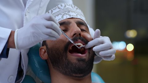 تصوير مقرب لطبيب يرتدي القفازات الطبية يقوم بفحص أسنان المريض وتشخيص المرض, العناية بالصحة الفموية، تركيب الحشوات ومعالجة التسوس، المعدات اللازمة لإجراء الفحوصات الطبية، شاب عربي خليجي عماني يرتدي الزي التقليدي يجلس على كرسي العلاج، زيارة العيادة السنية لإجراء الفحوصات الدورية