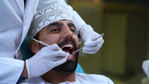 المعدات اللازمة لإجراء الفحوصات الطبية، شاب عربي خليجي عماني يرتدي الزي التقليدي يجلس على كرسي العلاج، تصوير مقرب لطبيب يرتدي القفازات الطبية يقوم بفحص أسنان المريض وتشخيص المرض, العناية بالصحة الفموية، تركيب الحشوات ومعالجة التسوس، زيارة العيادة السنية لإجراء الفحوصات الدورية