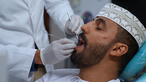 تصوير مقرب لطبيب يرتدي القفازات الطبية يقوم بفحص أسنان المريض وتشخيص المرض, العناية بالصحة الفموية، تركيب الحشوات ومعالجة التسوس، زيارة العيادة السنية لإجراء الفحوصات الدورية، شاب عربي خليجي عماني يرتدي الزي التقليدي يجلس على كرسي العلاج