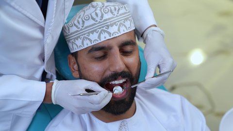 تركيب الحشوات ومعالجة التسوس، زيارة العيادة السنية لإجراء الفحوصات الدورية،العناية بالصحة الفموية، تصوير مقرب لطبيب يرتدي القفازات الطبية يقوم بفحص أسنان المريض وتشخيص المرض, شاب عربي خليجي عماني يرتدي الزي التقليدي يجلس على كرسي العلاج