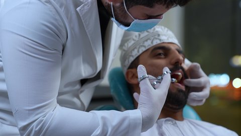 الاستعداد لحقن البنج الموضعي لتخفيف الشعور بالألم , يد طبيب أسنان يرتدي القفازات الطبية يمسك بيده إبرة المخدر، معالجة النخور والتسوس، تصوير مقرب لرجل عربي خليجي عماني يرتدي الزي التقليدي يجلس في العيادة السنية بإيماءات السرور, تقديم الرعاية الصحية في المراكز التخصصية