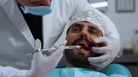معالجة النخور والتسوس، تصوير مقرب لرجل عربي خليجي عماني يرتدي الزي التقليدي يجلس في العيادة السنية بإيماءات السرور, تقديم الرعاية الصحية في المراكز التخصصية، الاستعداد لحقن البنج الموضعي لتخفيف الشعور بالألم , يد طبيب أسنان يرتدي القفازات الطبية يمسك بيده إبرة المخدر