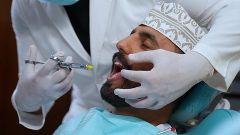 تقديم الرعاية الصحية في المراكز التخصصية، الاستعداد لحقن البنج الموضعي لتخفيف الشعور بالألم ,تصوير مقرب لرجل عربي خليجي عماني يرتدي الزي التقليدي يجلس في العيادة السنية بإيماءات السرور, يد طبيب أسنان يرتدي القفازات الطبية يمسك بيده إبرة المخدر،معالجة النخور والتسوس