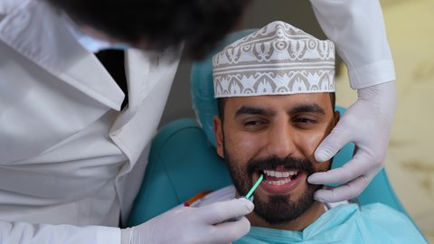 شاب عربي خليجي عماني يرتدي الزي التقليدي يجلس على كرسي العلاج ، تقديم الخدمات الطبية للمرضى, العناية بالصحة الفموية، تصوير مقرب لطبيب يرتدي القفازات يقوم بفحص أسنان المريض، مفهوم الطب والرعاية الصحية، استخدام الأدوات المعقمة لإجراء الفحوصات السنية