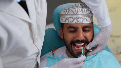 تقديم الخدمات الطبية للمرضى, العناية بالصحة الفموية، تصوير مقرب لطبيب يرتدي القفازات يقوم بفحص أسنان المريض، مفهوم الطب والرعاية الصحية، استخدام الأدوات المعقمة لإجراء الفحوصات السنية، شاب عربي خليجي عماني يرتدي الزي التقليدي يجلس على كرسي العلاج