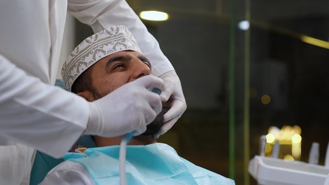 العناية بالصحة الفموية، تصوير مقرب لطبيب يرتدي القفازات يقوم بفحص أسنان المريض، مفهوم الطب والرعاية الصحية، استخدام الأدوات المعقمة لإجراء الفحوصات السنية، تقديم الخدمات الطبية للمرضى, شاب عربي خليجي عماني يرتدي الزي التقليدي يجلس على كرسي العلاج