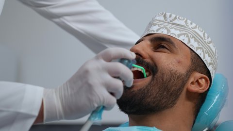 استخدام الأدوات المعقمة لإجراء الفحوصات السنية، العناية بالصحة الفموية، مفهوم الطب والرعاية الصحية، تقديم الخدمات الطبية للمرضى, تصوير مقرب لطبيب يرتدي القفازات يقوم بفحص أسنان المريض، شاب عربي خليجي عماني يرتدي الزي التقليدي يجلس على كرسي العلاج