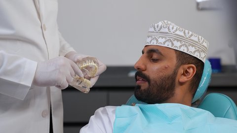 مفهوم الطب والرعاية الصحية, ايماءات الاطمئنان والسرور، الاهتمام في الصحة الفموية, تقديم نصائح للمحافظة على صحة الفم والأسنان، طبيب أسنان يرتدي المعطف و القفازات الطبية يمسك بيده نموذج سني، تصوير مقرب لرجل عربي خليجي عماني يرتدي الكمه يجلس على كرسي العلاج