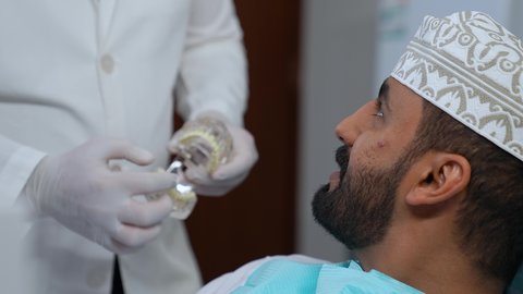 ايماءات الاطمئنان والسرور، الاهتمام في الصحة الفموية, تقديم نصائح للمحافظة على صحة الفم والأسنان،مفهوم الطب والرعاية الصحية, طبيب أسنان يرتدي المعطف و القفازات الطبية يمسك بيده نموذج سني، تصوير مقرب لرجل عربي خليجي عماني يرتدي الكمه يجلس على كرسي العلاج