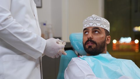 تقديم نصائح للمحافظة على صحة الفم والأسنان، ايماءات الاطمئنان والسرور، الاهتمام في الصحة الفموية, مفهوم الطب والرعاية الصحية, طبيب أسنان يرتدي المعطف و القفازات الطبية يمسك بيده نموذج سني، تصوير مقرب لرجل عربي خليجي عماني يرتدي الكمه يجلس على كرسي العلاج