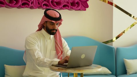 Gestures of integration and concentration during work, remote business management, browsing electronic applications and programs via laptop, using modern and advanced technical devices, a Saudi Arabian Gulf man wearing traditional thobe and shemagh working on a laptop in a cafe