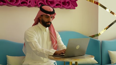 Remote business management, browsing electronic applications and programs via laptop, using modern and advanced technical devices, a Saudi Arabian Gulf man wearing traditional thobe and shemagh working on a laptop in a cafe