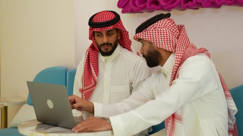 Dialogue and discussion between colleagues, using the laptop to complete projects and tasks in the cafe, spending quality time with friends, a close-up of a young Saudi Arabian Gulf man wearing a thobe and shemagh sitting with his friend in the cafe