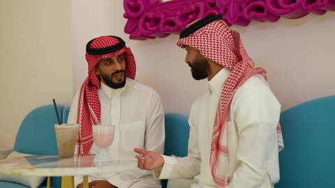 تبادل الاحاديث بين الأصدقاء، محاولة التخفيف من حرارة الصيف بالمشروب المنعش، ايماءات السعادة والسرور، تصويرمقرب لرجل عربي خليجي سعودي يرتدي الثوب و الشماغ يجلس فى احدي المقاهي برفقه صديقه