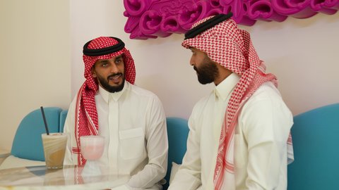 قضاء وقت ممتع في الخارج، محاولة التخفيف من حرارة الصيف بالمشروب المنعش، ايماءات السعادة والسرور، تصويرمقرب لرجل عربي خليجي سعودي يرتدي الثوب و الشماغ يجلس فى احدي المقاهي برفقه صديقه