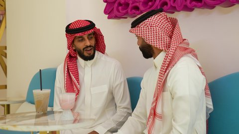 بناء علاقات اجتماعية ناجح، محاولة التخفيف من حرارة الصيف بالمشروب المنعش، ايماءات السعادة والسرور، تصويرمقرب لرجل عربي خليجي سعودي يرتدي الثوب و الشماغ يجلس فى احدي المقاهي برفقه صديقه