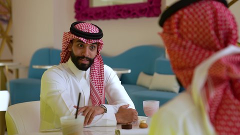 تبادل الحديث في احد المقاهي السعودية، محاولة التخفيف من حرارة الصيف بالمشروب المنعش، ايماءات السعادة والسرور، تصويرمقرب لرجل عربي خليجي سعودي يرتدي الثوب و الشماغ يجلس فى احدي المقاهي برفقه صديقه