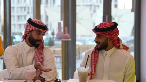 تبادل الأحاديث والأخبار، ايماءات السعادة والسرور اثناء تناول المشروبات الباردة، قضاء أوقات ممتعة في الويكند، الترويح عن النفسة برفقة الأصدقاء، تصوير لرجل عربي خليجي سعودي يرتدي الثوب و الشماغ يجلس فى المقهي برفقة صديقة