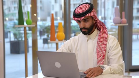 تعابير الاندماج والتركيز أثناء العمل عبر الحاسب المحمول، الجلوس في إحدى المقاهي الراقية، استخدام الأجهزة التكنولوجية الحديثة والمتطورة، رجل عربي خليجي سعودي يرتدي الشماغ والثوب التقليدي يجلس في الكافية يستخدم اللابتوب