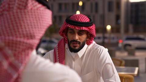 الاستمتاع بتناول المشروبات، قضاء أوقات ممتعة في الويكند، الترويح عن النفسة برفقة الأصدقاء، تصوير مقرب لرجل عربي خليجي سعودي يرتدي الثوب و الشماغ يجلس فى المقهي برفقة صديقة، ايماءات السعادة و السرور