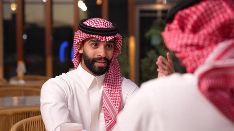 قضاء أوقات ممتعة في الويكند، الترويح عن النفسة برفقة الأصدقاء، الاستمتاع بتناول المشروبات ، تصوير مقرب لرجل عربي خليجي سعودي يرتدي الثوب و الشماغ يجلس فى المقهي برفقة صديقة، ايماءات السعادة و السرور