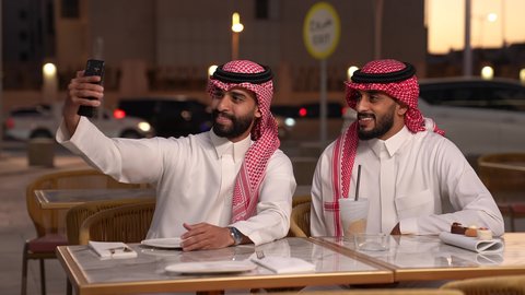اجراء مكالمة فيديو، صورة جماعية ، توثيق اللحظات السعيدة ، بث مباشر، تصوير مقرب لرجل عربي خليجي سعودي يرتدي الزي التقليدي يجلس في المقهي برفقه صديقة و يقوم بالتقاط صورة سيلفي عبر الهاتف المحمول بايماءات السعادة و السرور