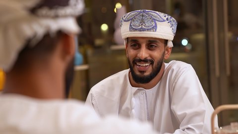 حوار ونقاش مع الاصدقاء، علاقات إجتماعية بين الإصدقاء، قضاء وقت ممتع ومسلي في احدى الكافيهات، تصوير مقرب لرجل عربي خليجي عماني يرتدي الدشداشة و الكمه يجلس فى المقهي برفقة صديقة ، الاستمتاع بتناول المشروبات