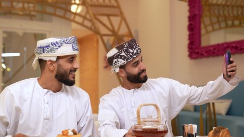 استخدام التقنيات الحديثة والمتطورة،  إجراء مكالمة فيديو ، التقاط صور سيلفي تذكارية باستخدام الهاتف المحمول، تصوير مقرب لرجل عربي خليجي عماني يرتدي الدشداشة و الكمه يقوم بامساك الجوال برفقه صديقه،  توثيق اللحظات السعيدة