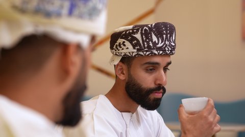 جلسة وناسة وتعارف، علاقات إجتماعية بين الإصدقاء، قضاء وقت ممتع ومسلي في احدى الكافيهات، تصوير مقرب لرجل عربي خليجي عماني يرتدي الدشداشة و الكمه يجلس فى المقهي برفقة صديقة و يقوم بشرب فنجان من القهوه بايماءات السعادة و السرور