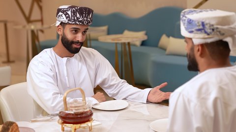 قضاء وقت ممتع في الخارج، حوار بين اصدقاء في مطعم، تصوير مقرب لرجل عربي خليجي عماني يرتدي الدشداشة و الكمه يقوم بتناول قطعه الحلويات برفقة صديقة، التعارف وبناء علاقات اجتماعيه, صداقات ومعارف