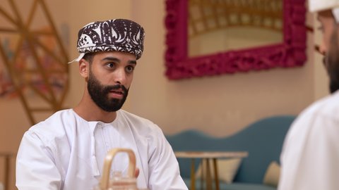 التعارف وبناء علاقات اجتماعيه، حوار بين اصدقاء في مطعم، تصوير مقرب لرجل عربي خليجي عماني يرتدي الدشداشة و الكمه يجلس يرفقه صديقة في المقهى, مناقشة مهام العمل في المطعم, صداقات ومعارف، الدفع والشراء من خلال برامج وتطبيقات الجوال، الاستمتاع بتناول المشروبات