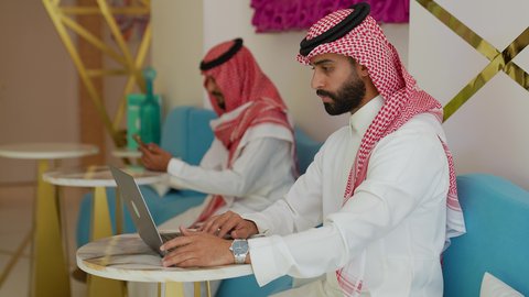 Performing office tasks and work, a side view of a Saudi Arabian Gulf man wearing traditional thobe sitting inside a cafe working on a laptop, integrating technology into practical life, working in a cafe or restaurant in the Kingdom of Saudi Arabia
