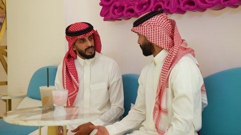 مشاركة الأصدقاء الأوقات الممتعة، ايماءات السعاده و السرور، الاستمتاع باجواء هادئه خارج المنزل، الترفيه والتسلية في العطلة، تصوير مقرب لرجل عربي خليجي سعودي يرتدي الثوب و الشماغ يجلس في احدي المقاهي برفقه صديقه