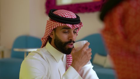 حوار ونقاش مع الاصدقاء، الاستمتاع باجواء هادئه خارج المنزل، تناول المشروبات الباردة،الترفيه والتسلية في العطلة، تصوير مقرب لرجل عربي خليجي سعودي يرتدي الثوب و الشماغ يجلس في احدي المقاهي و يمسك بيده كوب من المشروب
