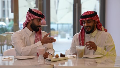 Fun and entertainment on vacation, chatting with friends, having a good time in a cafe, a close-up of a Saudi Arabian Gulf man wearing a thobe and shemagh sitting with his friend in a cafe with gestures of happiness and pleasure, enjoying a drink
