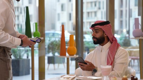 Entertainment and leisure on vacation, payment and purchase through mobile programs and applications, close-up of a Saudi Arabian Gulf man wearing a thobe and shemagh sitting in a cafe, spending a pleasant time outside the home