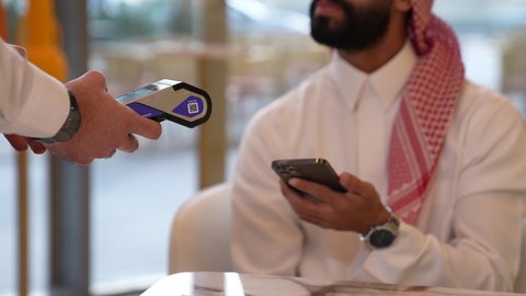 Payment and purchase through mobile programs and applications, a close-up of a Saudi Arabian Gulf man wearing a thobe and a shemagh sitting in a cafe, spending a fun time outside the home, entertainment and recreation on vacation
