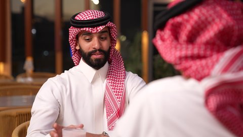 قضاء وقت ممتع في الخارج، جلسة وناسة وتعارف، ايماءات السعادة و السرور، الاستمتاع بتناول المشروبات ، تصوير مقرب لرجل عربي خليجي سعودي يرتدي الثوب و الشماغ يجلس فى المقهي برفقة صديقة
