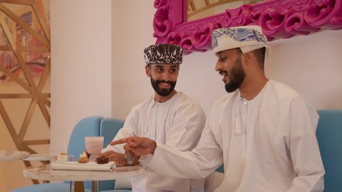 A pleasant and relaxing session, gestures of happiness and pleasure, enjoying food and drinks, spending a fun and entertaining time in a café, a close-up of an Omani Gulf Arab man wearing a dishdasha and a kummah sitting in a café with his girlfriend