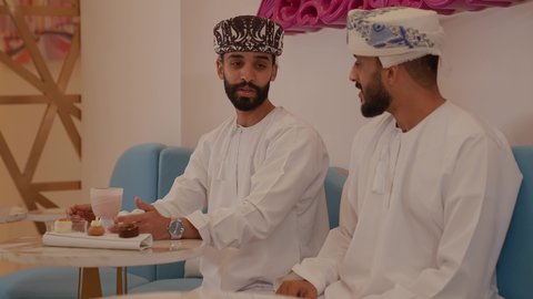 ايماءات السعادة و السرور، الاستمتاع بتناول الماكولات و المشروبات ، قضاء وقت ممتع ومسلي في احدى الكافيهات، جلسة وناسة وتعارف، تصوير مقرب لرجل عربي خليجي عماني يرتدي الدشداشة و الكمه يجلس فى المقهي برفقة صديقة