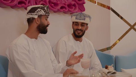 الاستمتاع بتناول الماكولات و المشروبات ، ايماءات السعادة و السرور، قضاء وقت ممتع ومسلي في احدى الكافيهات، جلسة وناسة وتعارف، تصوير مقرب لرجل عربي خليجي عماني يرتدي الدشداشة و الكمه يجلس فى المقهي برفقة صديقة