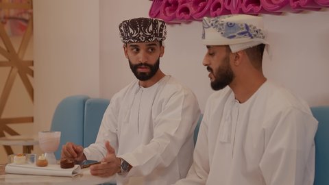 The concept of documenting happy moments, spending a fun and entertaining time in a café, a fun and friendly session, a close-up of an Arab Gulf Omani man wearing a dishdasha and a kuma sitting in a café with his girlfriend, taking souvenir selfies using his mobile phone