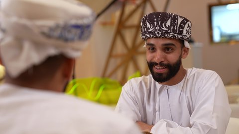 علاقات إجتماعية بين الإصدقاء، قضاء وقت ممتع ومسلي في احدى الكافيهات، جلسة وناسة وتعارف، تصوير مقرب لرجل عربي خليجي عماني يرتدي الدشداشة و الكمه يجلس فى المقهي برفقة صديقة بايماءات السعادة و السرور