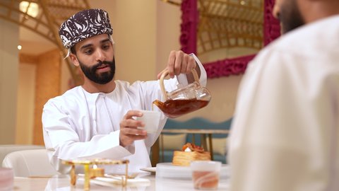 الاستمتاع بشرب المشروبات الساخنة في احدى الكافيهات، حوار بين اصدقاء في مطعم، تصوير مقرب لرجل عربي خليجي عماني يرتدي الدشداشة و الكمه يجلس برفقه صديقة في المقهى يقوم بصب الشاي، التعارف وبناء علاقات اجتماعيه, صداقات ومعارف
