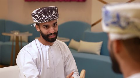 Payment and purchase through mobile programs and applications, a conversation between friends in a restaurant, a close-up of an Arab Gulf Omani man wearing a dishdasha and a kuma sitting with his girlfriend in a cafe, discussing work tasks in the restaurant, getting to know each other and building social relationships, friendships and acquaintances